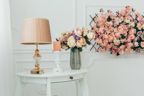 White bedside table with drawers, a lamp and flowers beside a bed